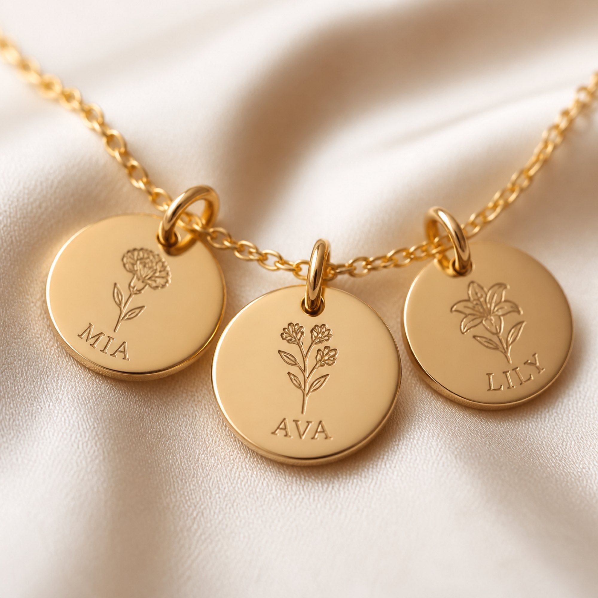 Close-up of a gold necklace with three engraved discs showing birth flowers and names, on cream silk