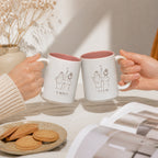 Two hands clinking the matching printed mugs over a cream linen table with a plate of biscuits
