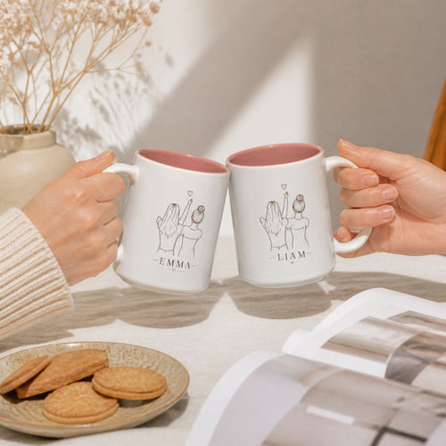Two hands clinking the matching printed mugs over a cream linen table with a plate of biscuits