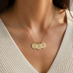 A gold three-disc engraved name necklace worn against a cream ribbed knit top
