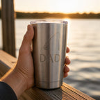 A hand holding an engraved stainless tumbler on a dock rail beside a lake at golden hour